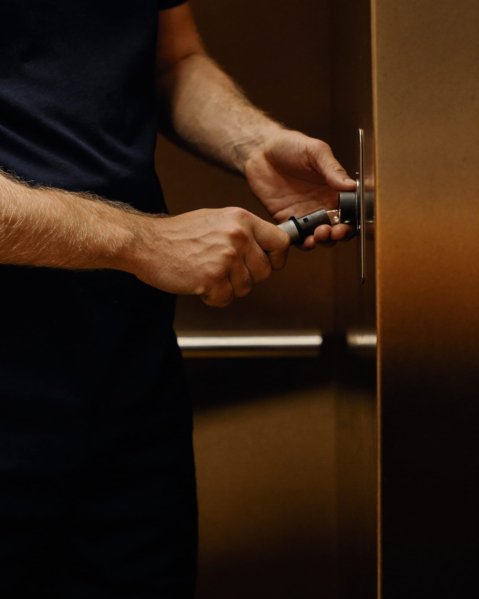 Mani di un tecnico ascensorista che intervengono con un attrezzo di precisione sul meccanismo di una porta di piano in acciaio, nella luce calda della cabina